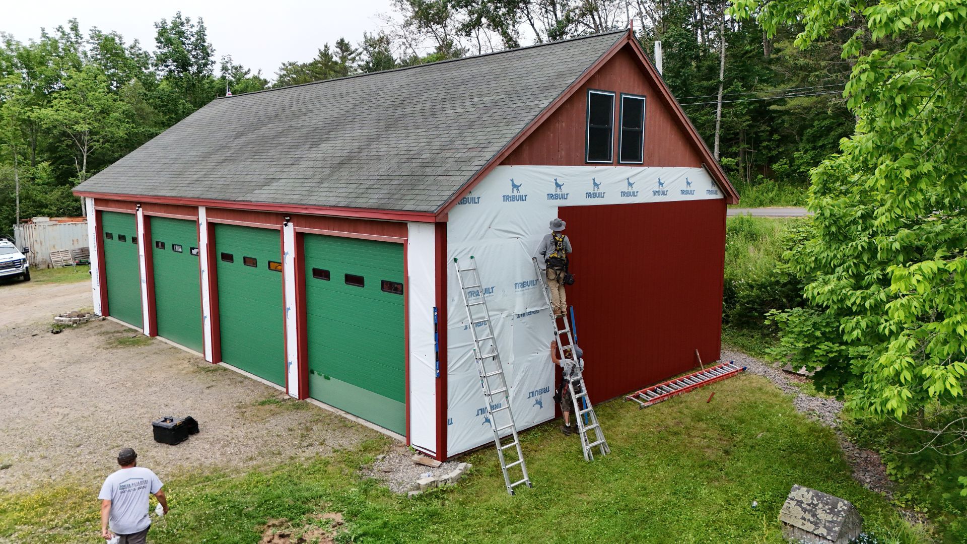 Three-bay garage under renovation; worker on ladder, red siding, green garage doors, surrounded by trees.