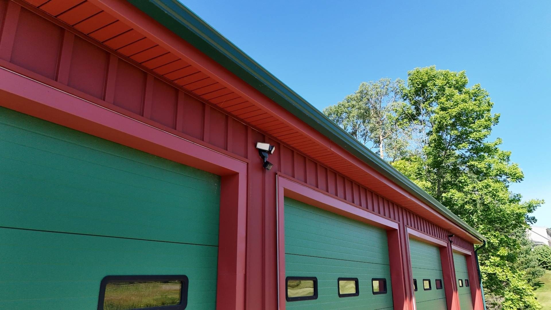 Red and green building with three garage doors, security light, and trees against blue sky.