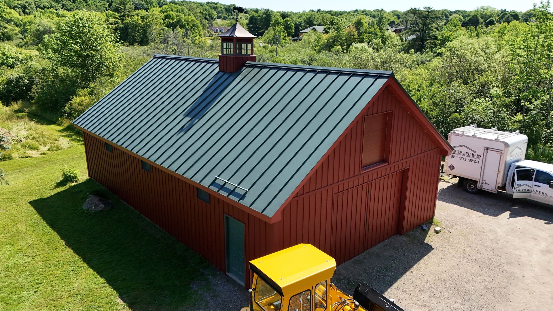 Red barn with green metal roof, cupola, and yellow tractor in front; vehicles parked alongside.