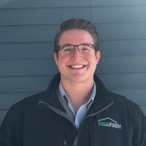 Man in glasses, smiling, wearing a black jacket and button-down shirt, in front of blue siding.