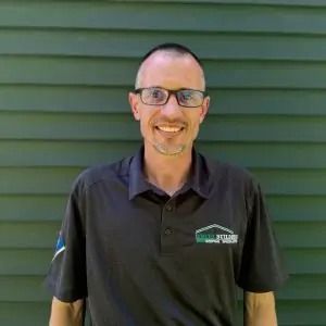 Man with glasses smiles in front of a green wall. He wears a dark shirt with a logo.