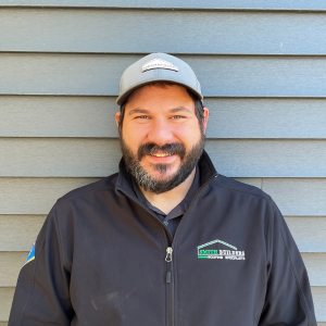 Man with beard, gray cap and black jacket, smiling in front of gray siding.