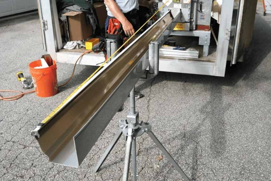 A person measures a metal gutter on a tripod stand; tools and truck are in the background.