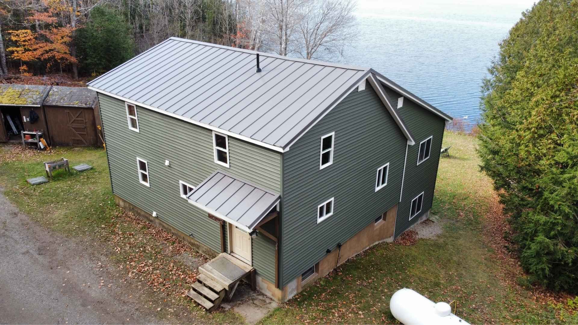 Green house with metal roof near a lake, surrounded by trees and foliage.