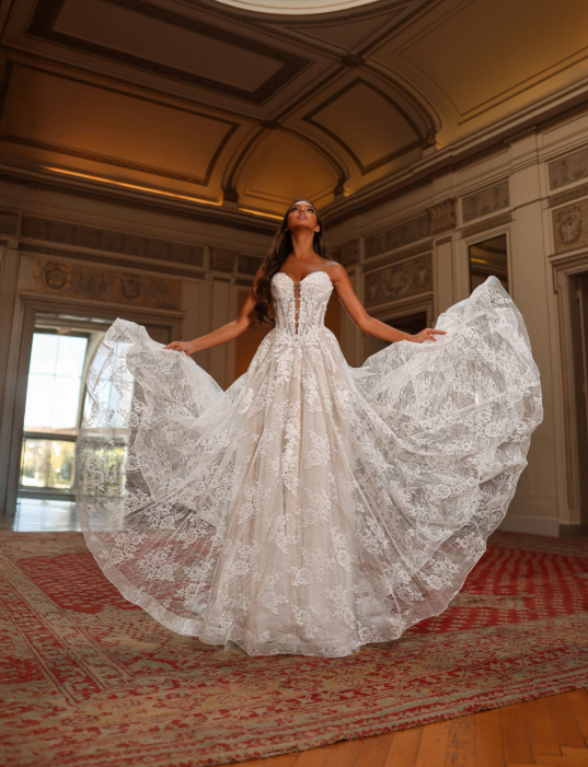 Woman in a white lace wedding gown, arms outstretched, indoors.