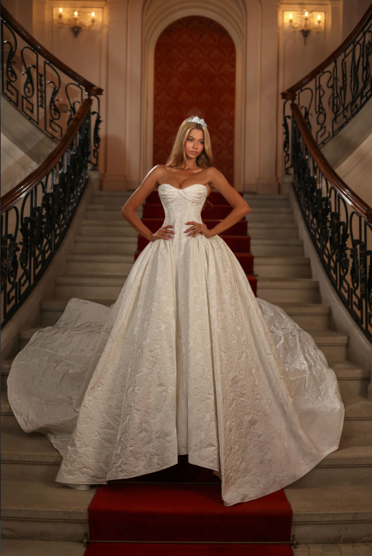 Woman in white ballgown poses on a grand staircase with red carpet; tiara.