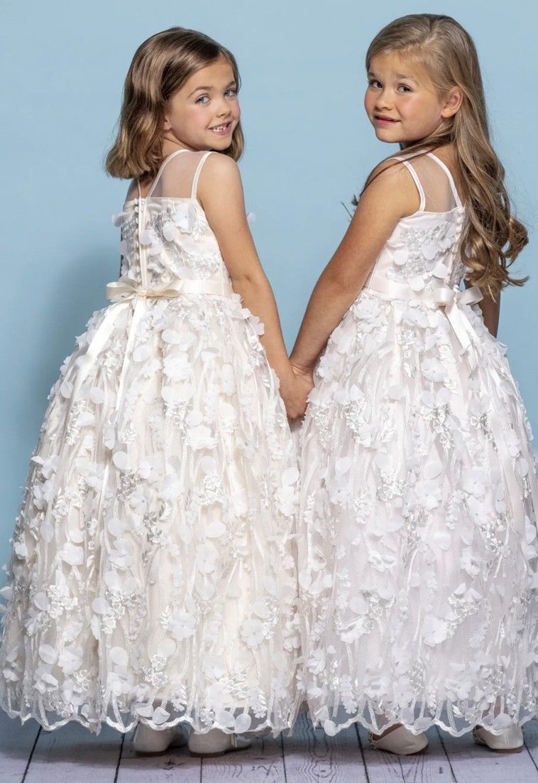 Two girls in white floral dresses holding hands, smiling against a light blue backdrop.