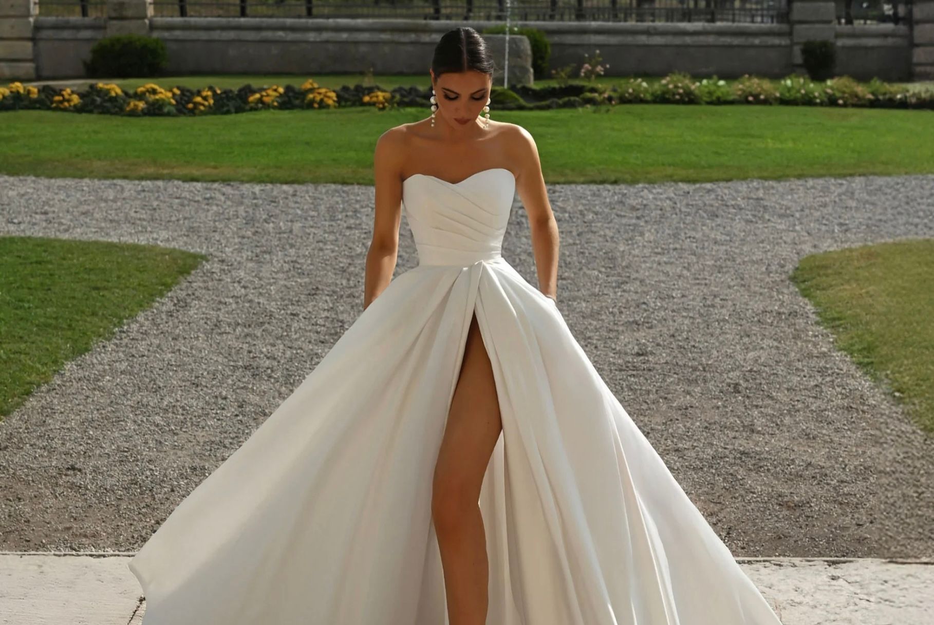 Woman in strapless white gown with thigh-high slit, posing outside on stone pathway and grass.