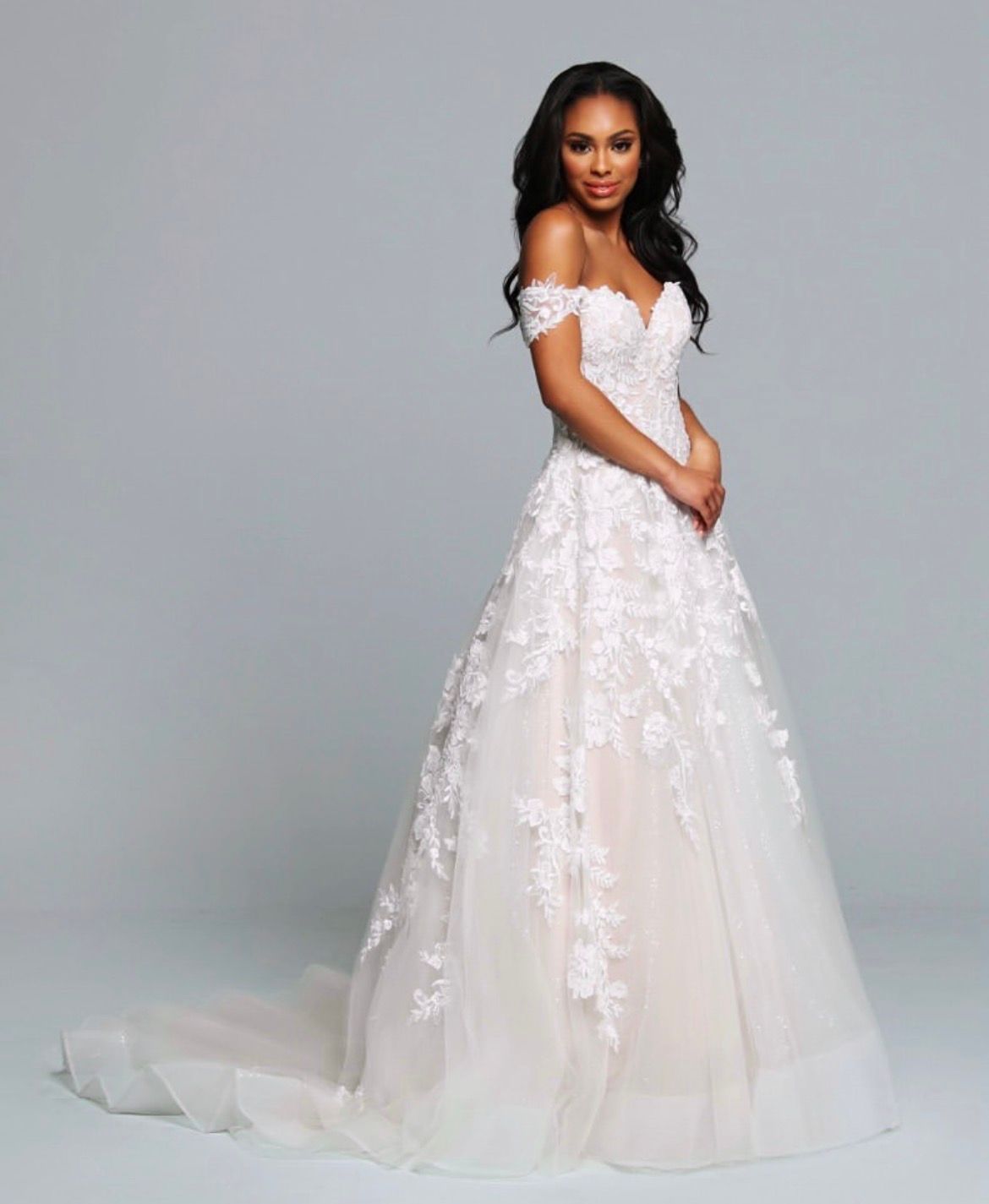 Woman in a white, mermaid-style wedding dress with lace details and a train, posing against a gray backdrop.