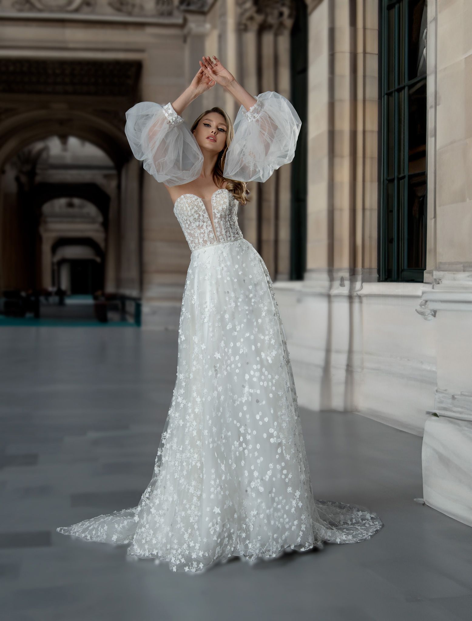 Woman in a white floral wedding gown with puffy sleeves, arms raised, in a stone hallway.