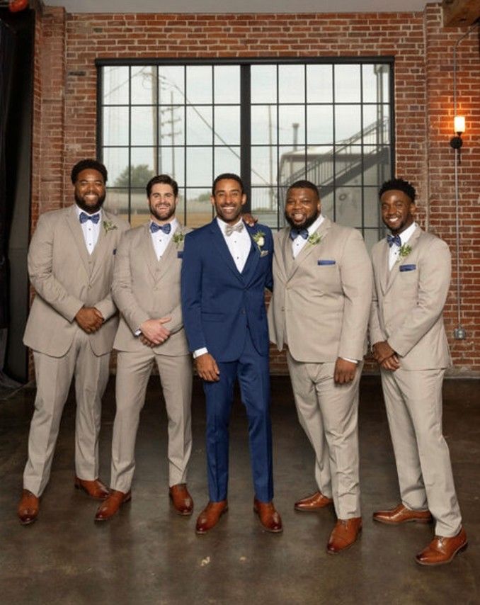 Five men in suits pose for a photo in front of a window. Groom wears navy, groomsmen wear tan suits.