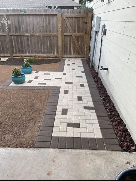 Brick pathway in a backyard, bordered by brown pavers and gravel. Blue planters sit near the pathway.