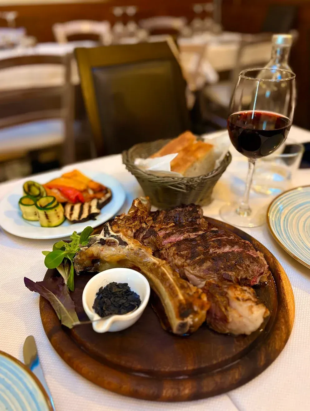 Bistecca alla griglia su un tagliere di legno con verdure, pane e vino rosso in un ambiente da ristorante.
