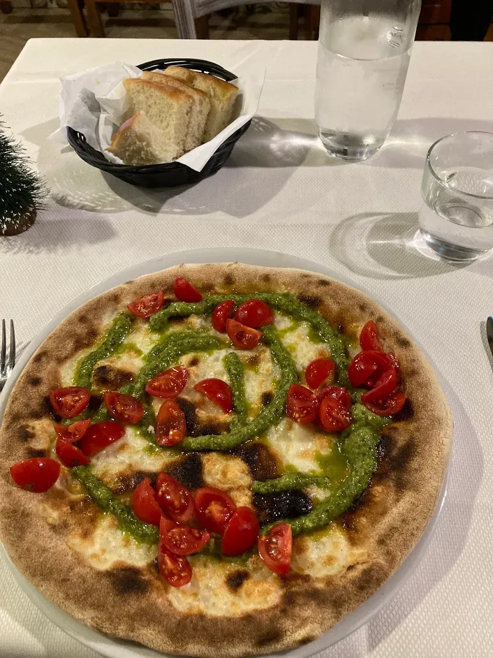 Pizza con pesto verde e pomodorini su un tavolo bianco, con un bicchiere d'acqua e del pane sullo sfondo.