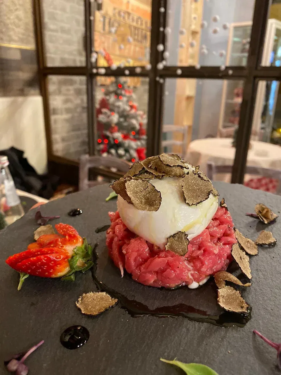 Dessert impiattato con mousse rosa, crema bianca, guarnizione al cioccolato e fragola su un piatto di ardesia nera.