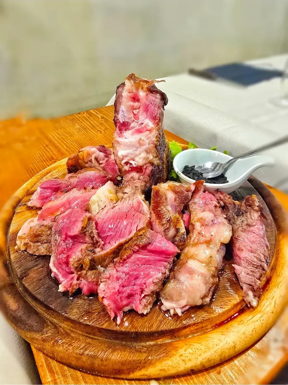 Una bistecca tagliata a fette sottili, cotta al sangue, servita su un piatto di legno con una ciotolina di salsa e un cucchiaio.