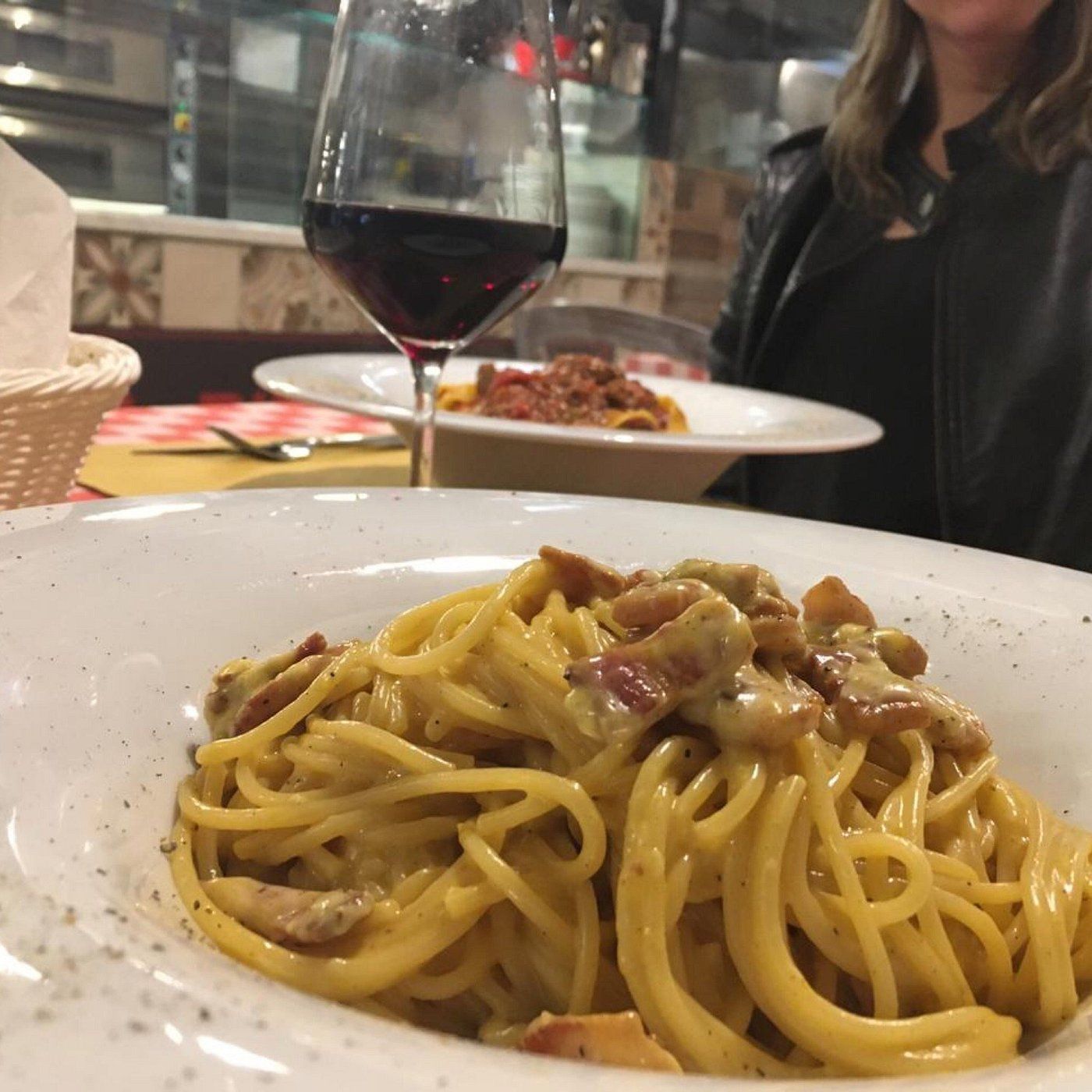 In primo piano un piatto di spaghetti con un bicchiere di vino rosso e un altro piatto di pasta su un tavolo di un ristorante.