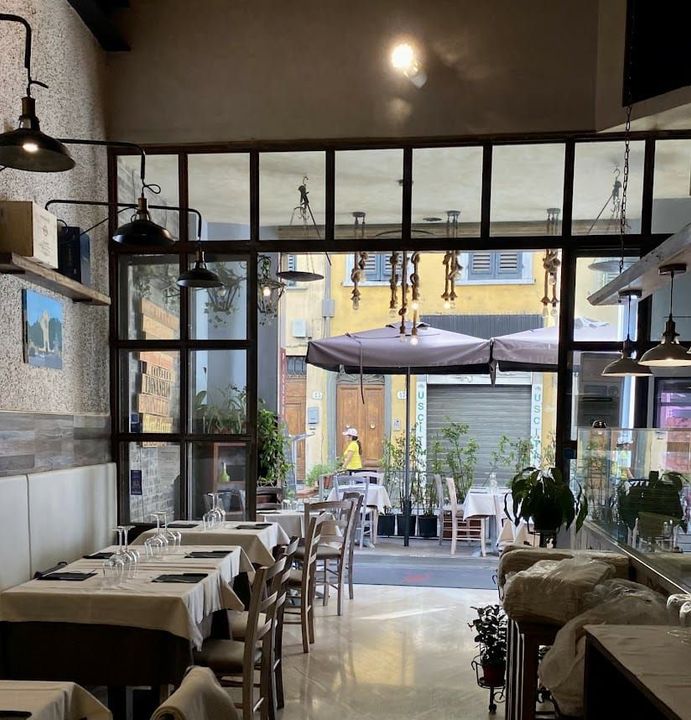 Interni moderni di un caffè con lunghi tavoli, lampade a sospensione e vista su un patio esterno attraverso ampie vetrate.