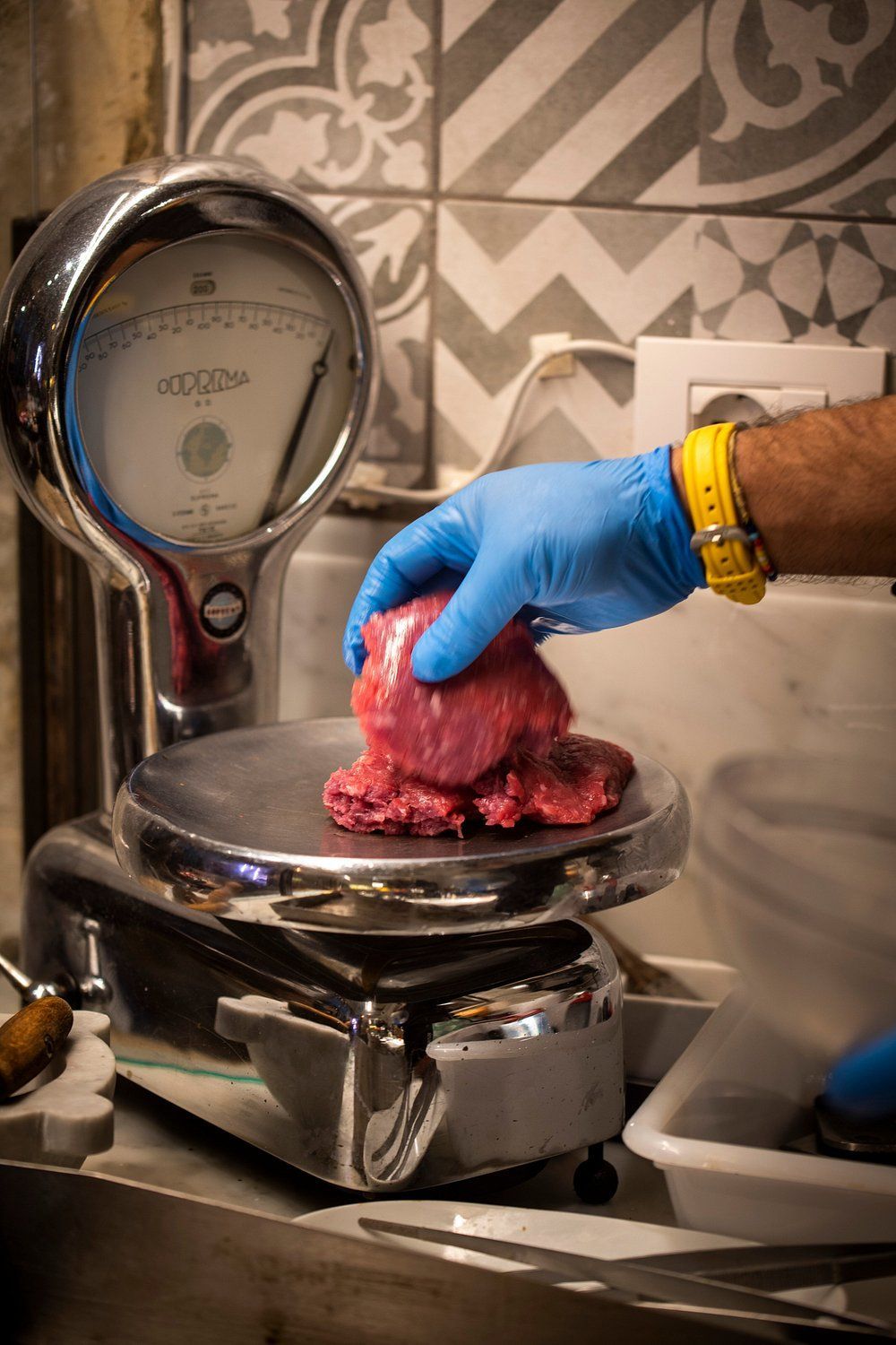 Mano guantata che posiziona carne cruda su una bilancia da cucina accanto a un lavandino.