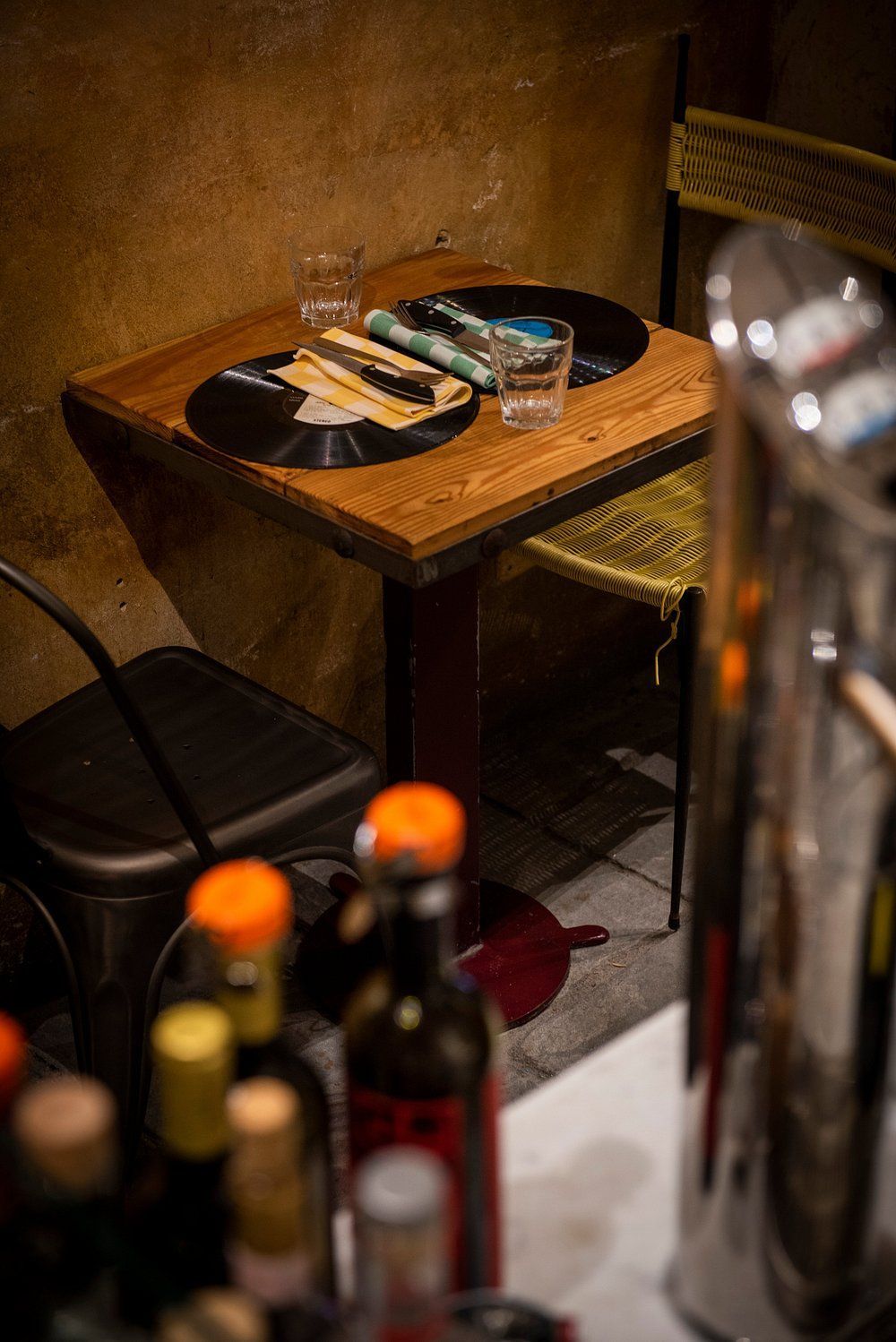 Un piccolo tavolo da ristorante con un bicchiere, piatti, bacchette e bottiglie in primo piano.