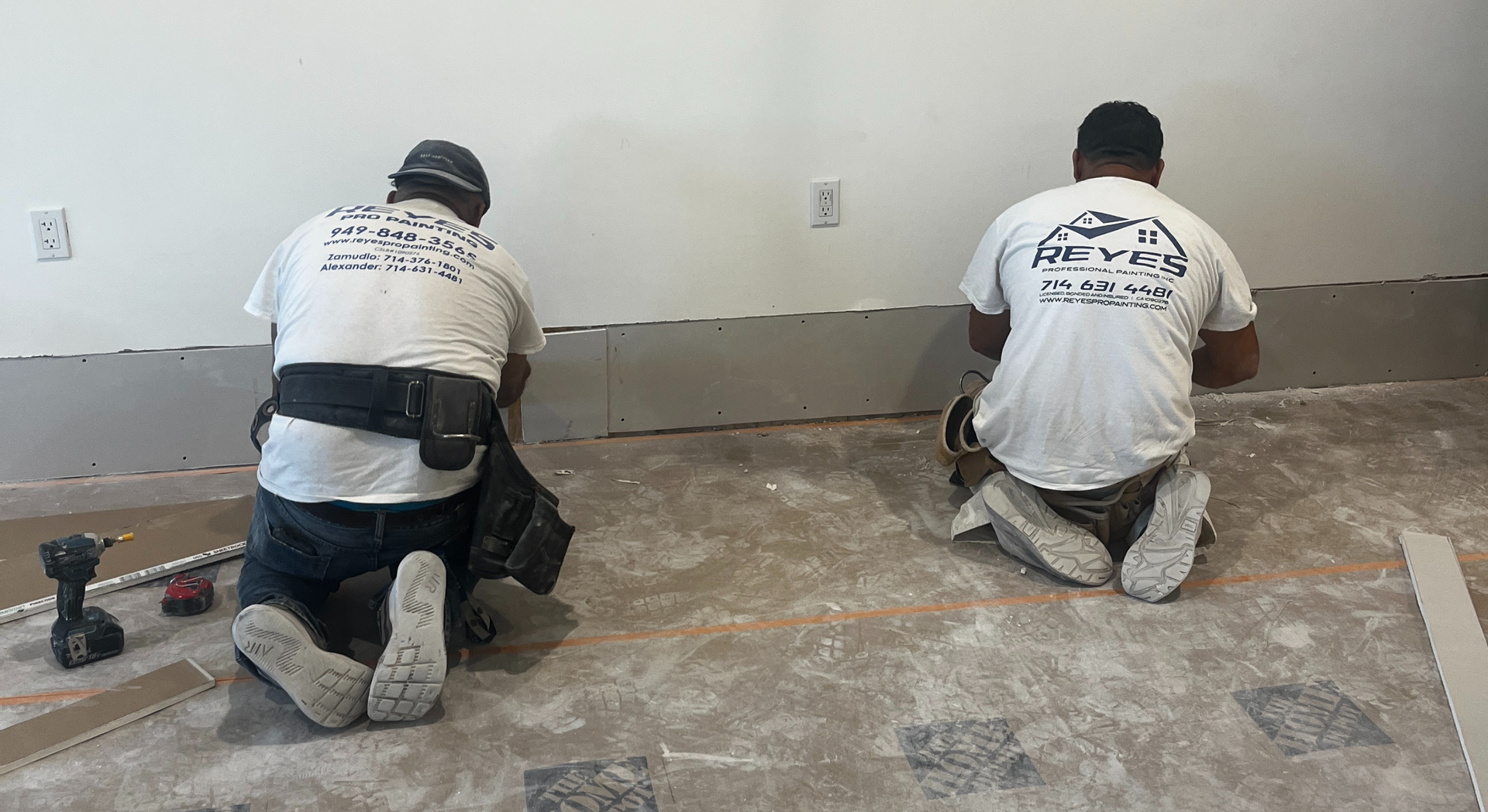 Two workers kneeling, installing flooring along a wall. They wear work clothes and have tools.