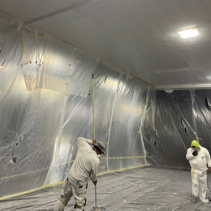 Two workers in protective suits mixing paint in a plastic-covered room.