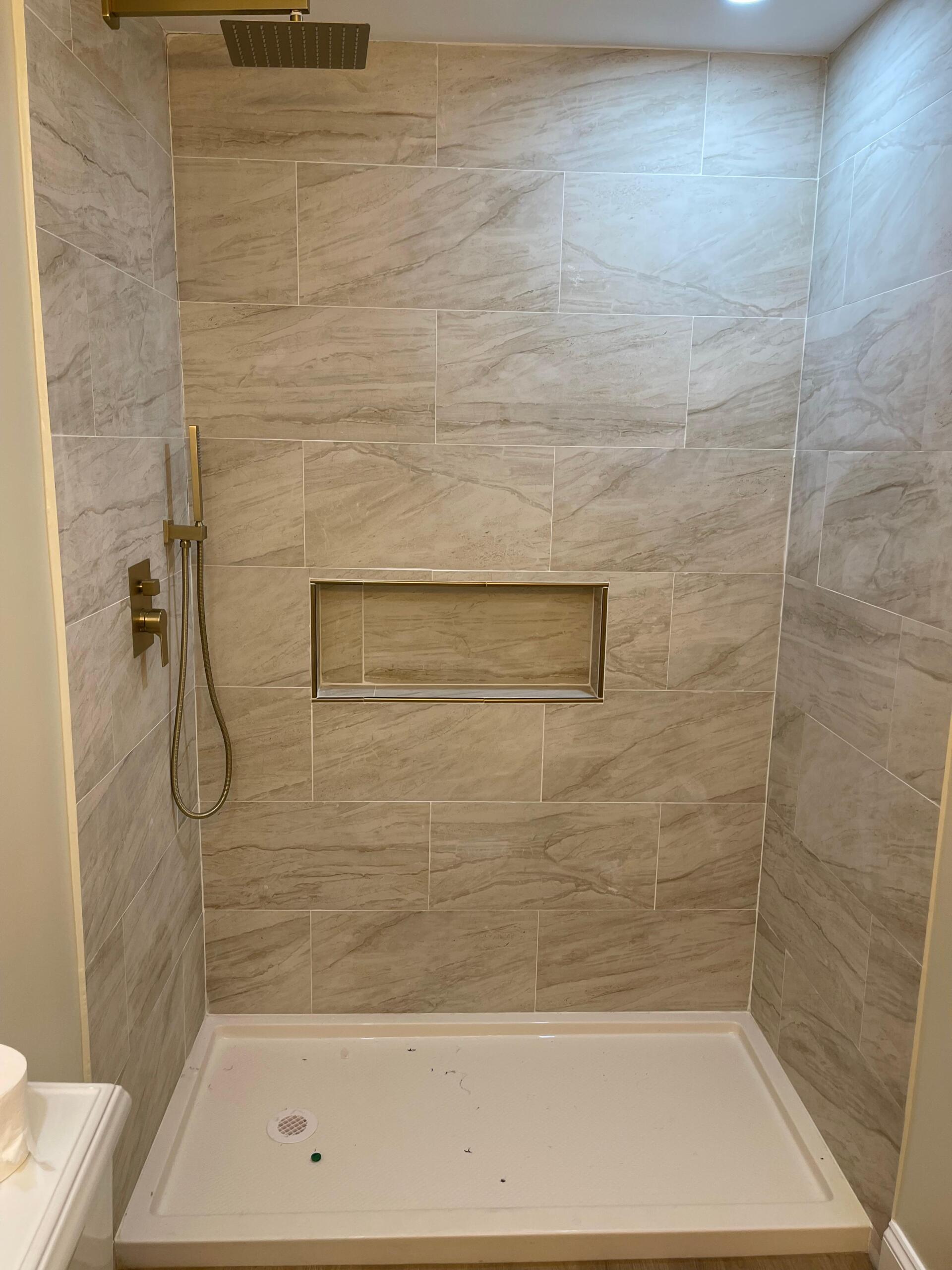 Newly tiled shower with a white base, gold showerhead, and a recessed shelf.