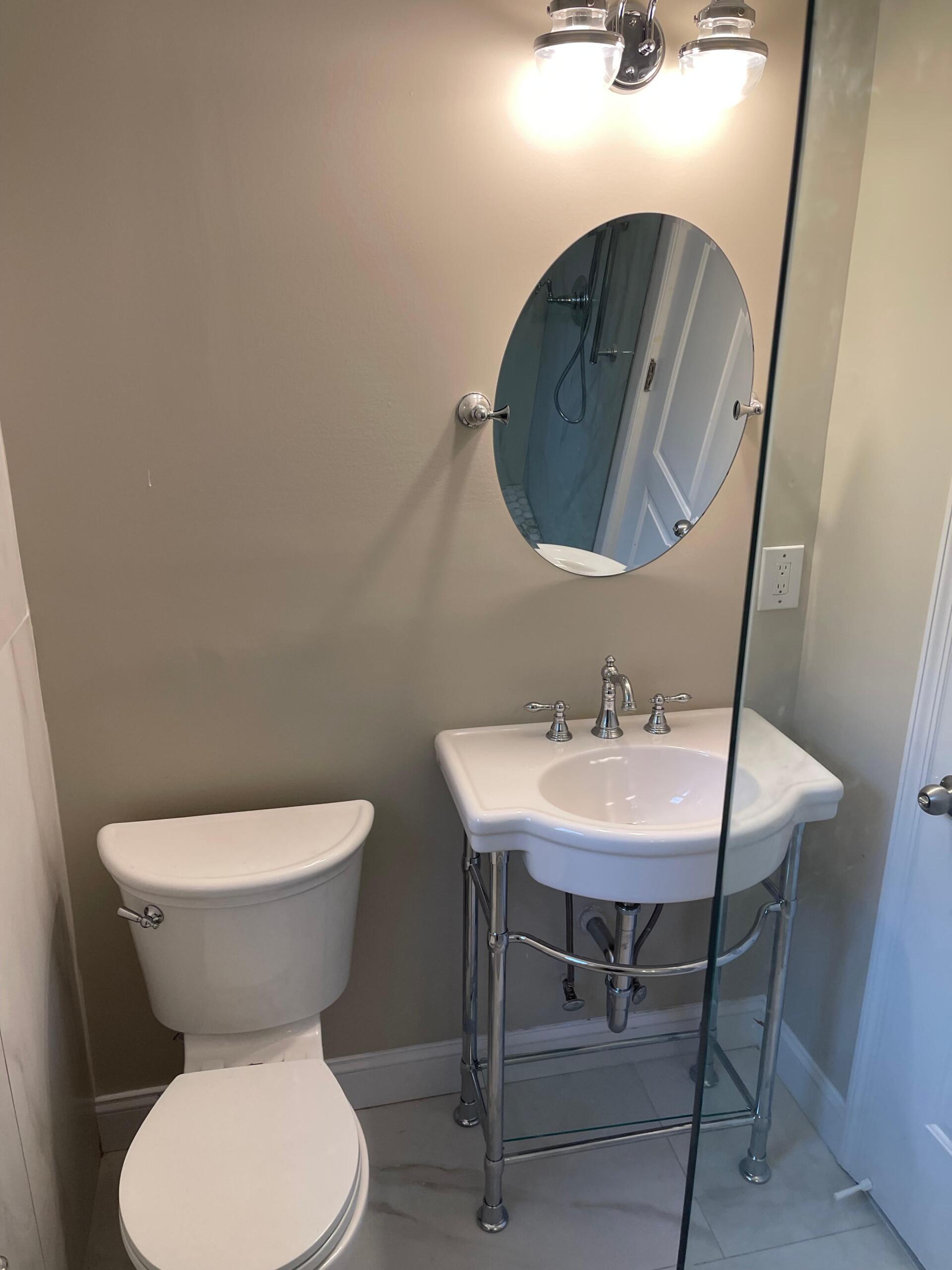 Bathroom with toilet, sink, mirror, and overhead lighting. Beige walls, white fixtures, and chrome accents.