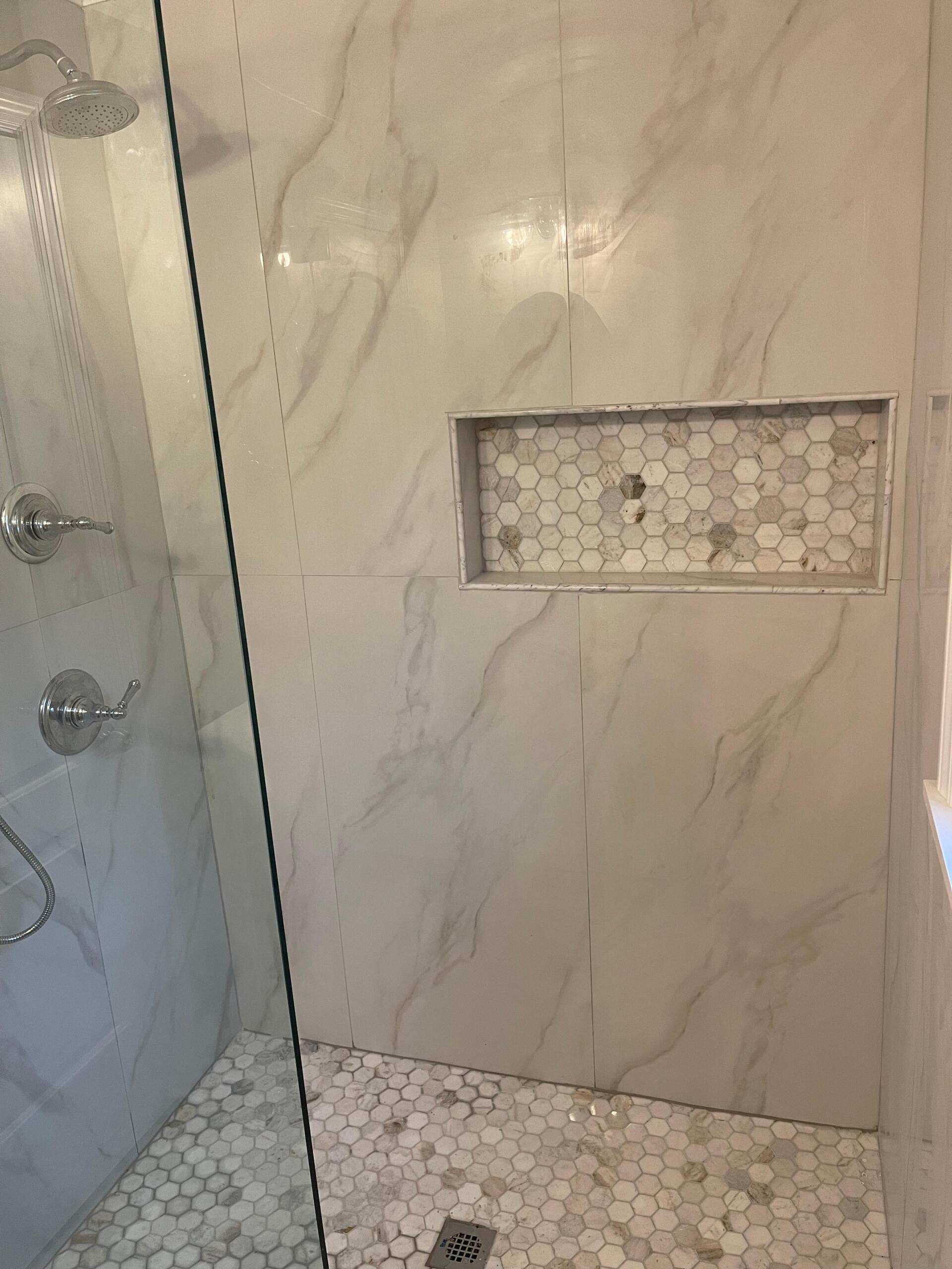 Shower interior with marble-patterned tile walls, mosaic floor, built-in shelf, glass door, and chrome fixtures.