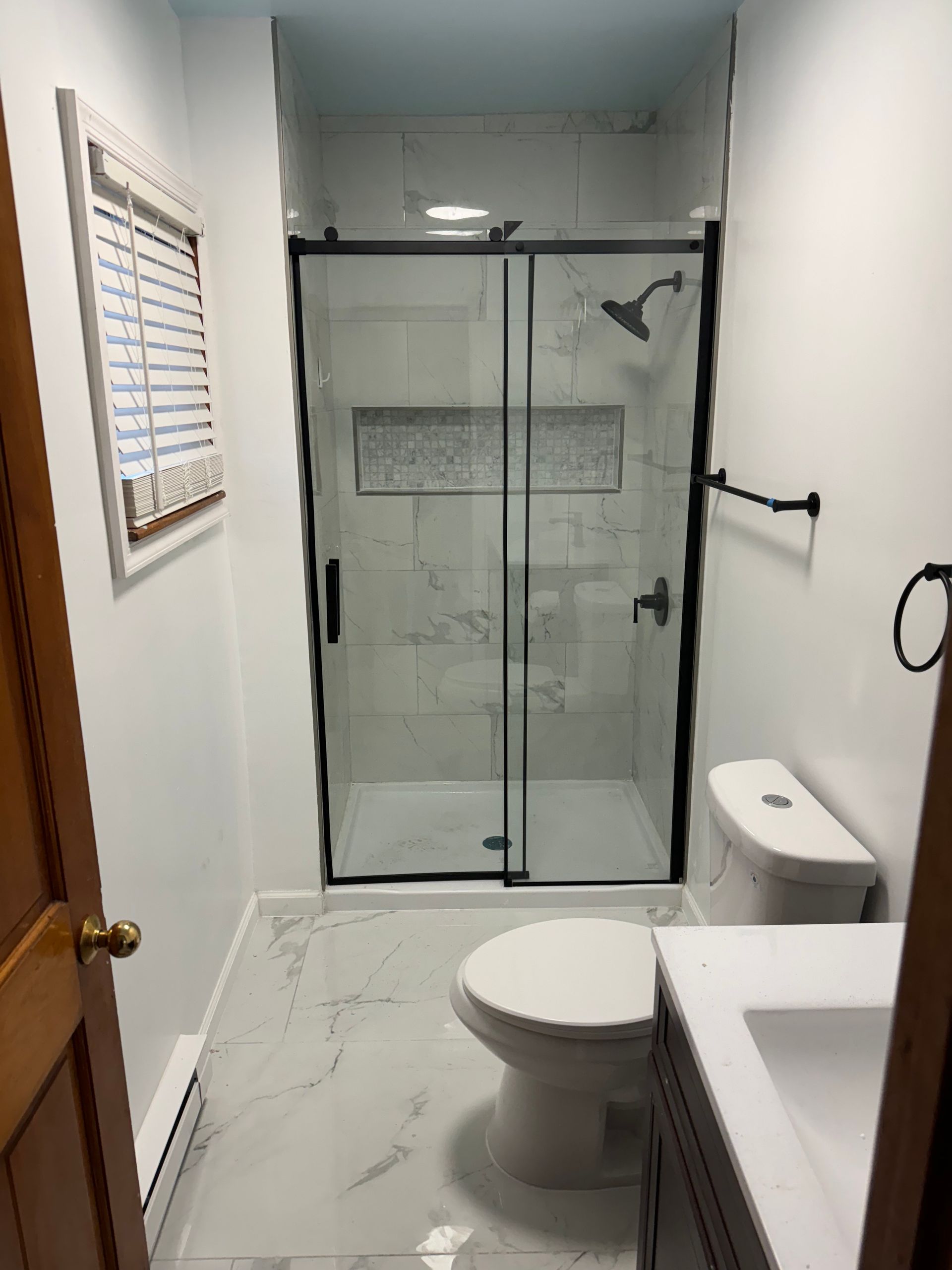 Bathroom with a shower, toilet, and sink. White walls, grey marble-look floor tiles, black shower frame, and hardware.