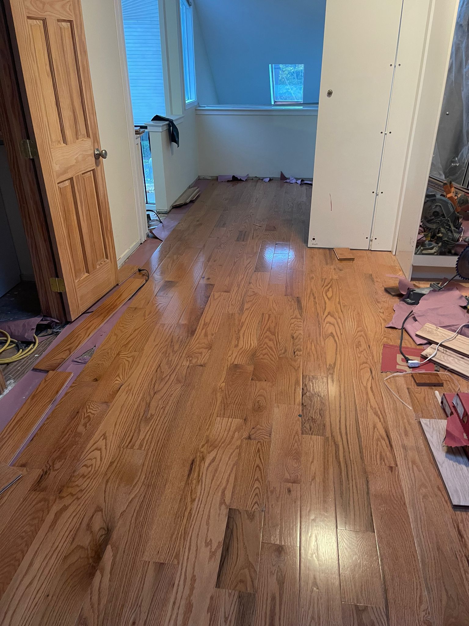 Wood flooring installation in progress; hallway with unfinished boards, door, and stairs.