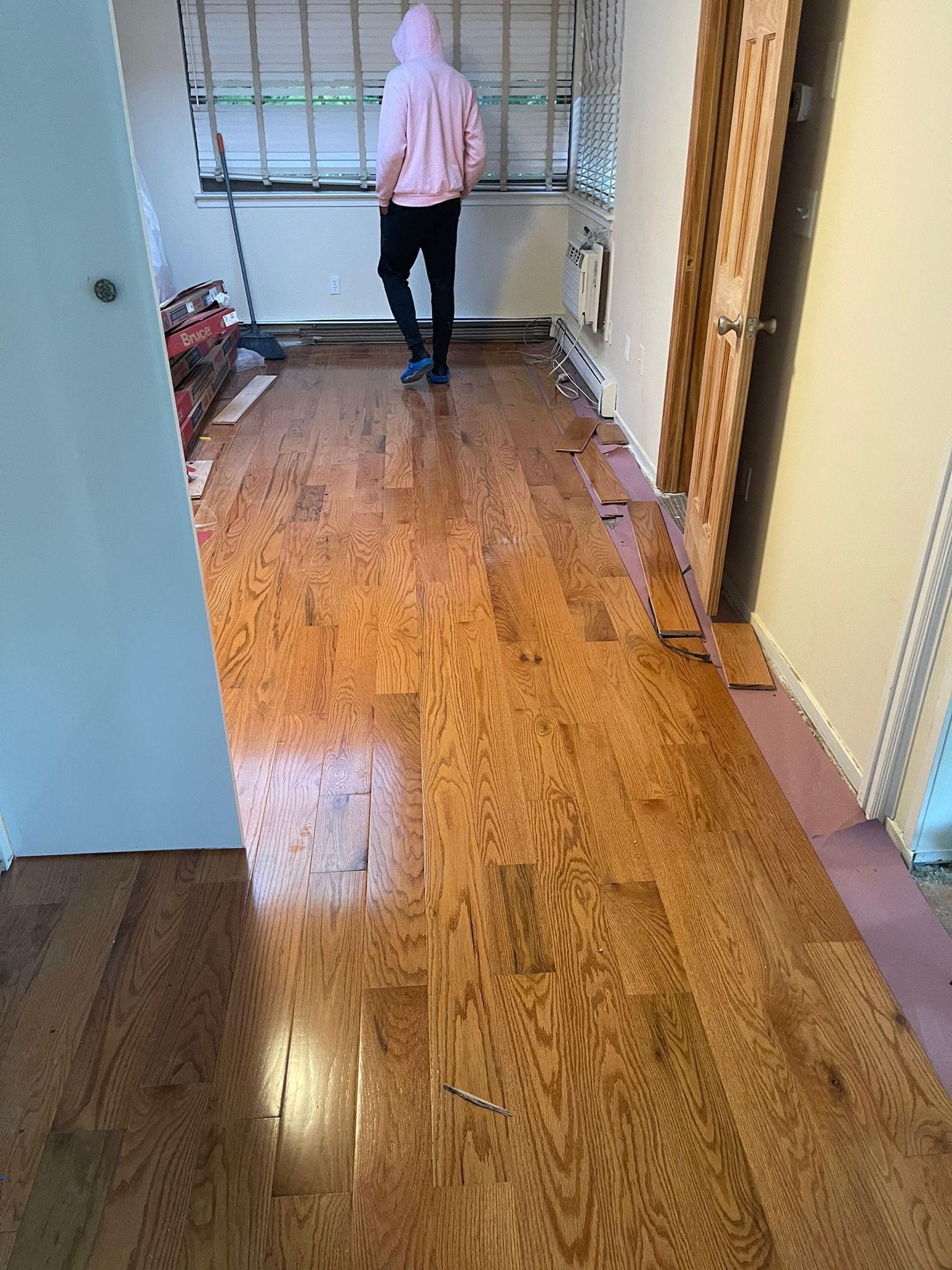 Person in pink hoodie walks on newly installed hardwood floor, doorway on the right.