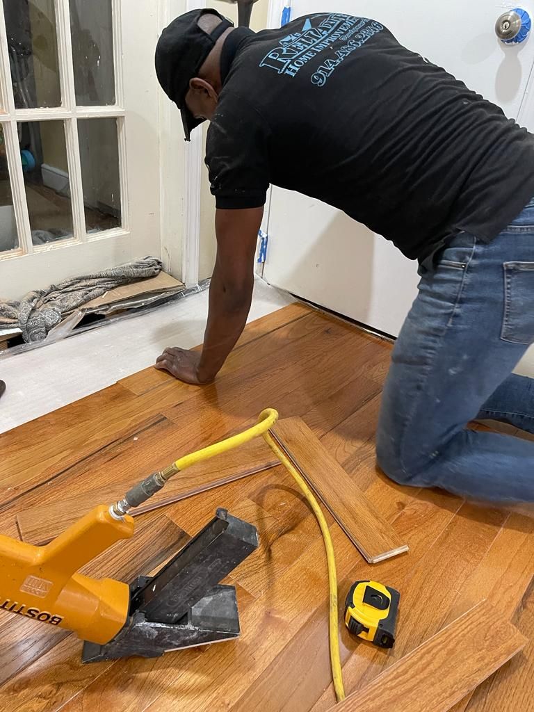 Person installing hardwood floor. Kneeling, using a yellow pneumatic nailer. Door and window in background.