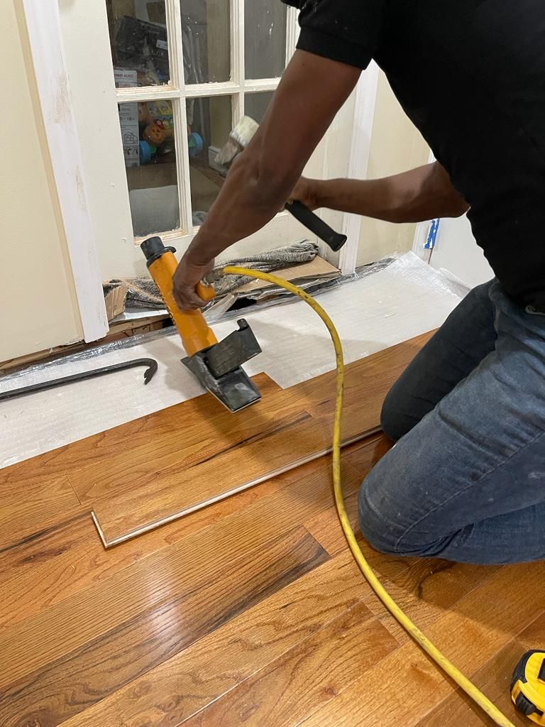 Person kneeling, installing hardwood flooring with a pneumatic nailer near a window. Yellow hose attached.