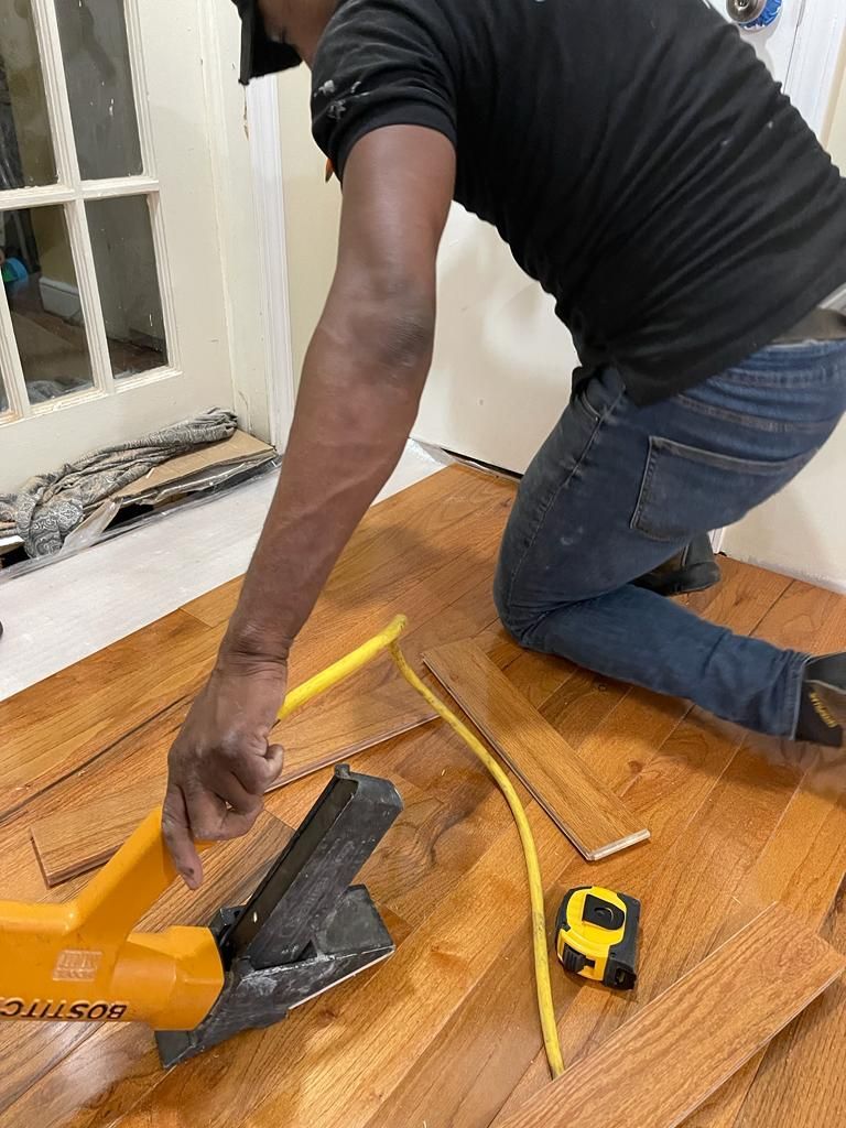 Person kneeling, cutting wood flooring with a yellow tool. Wood scraps, measuring tape, and wiring present.
