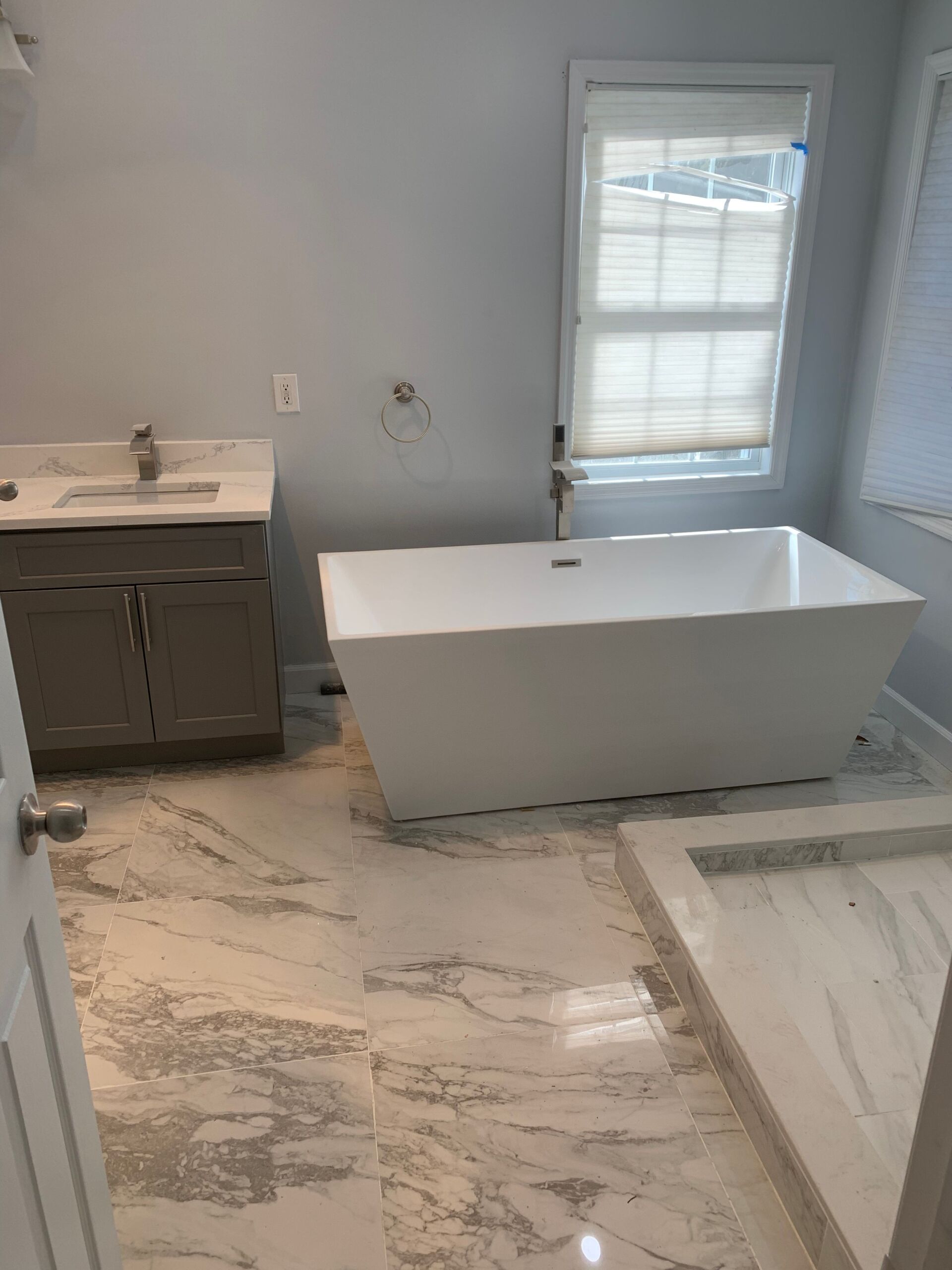 Bathroom with white rectangular tub, gray vanity, marble-look tile, and window with blinds.