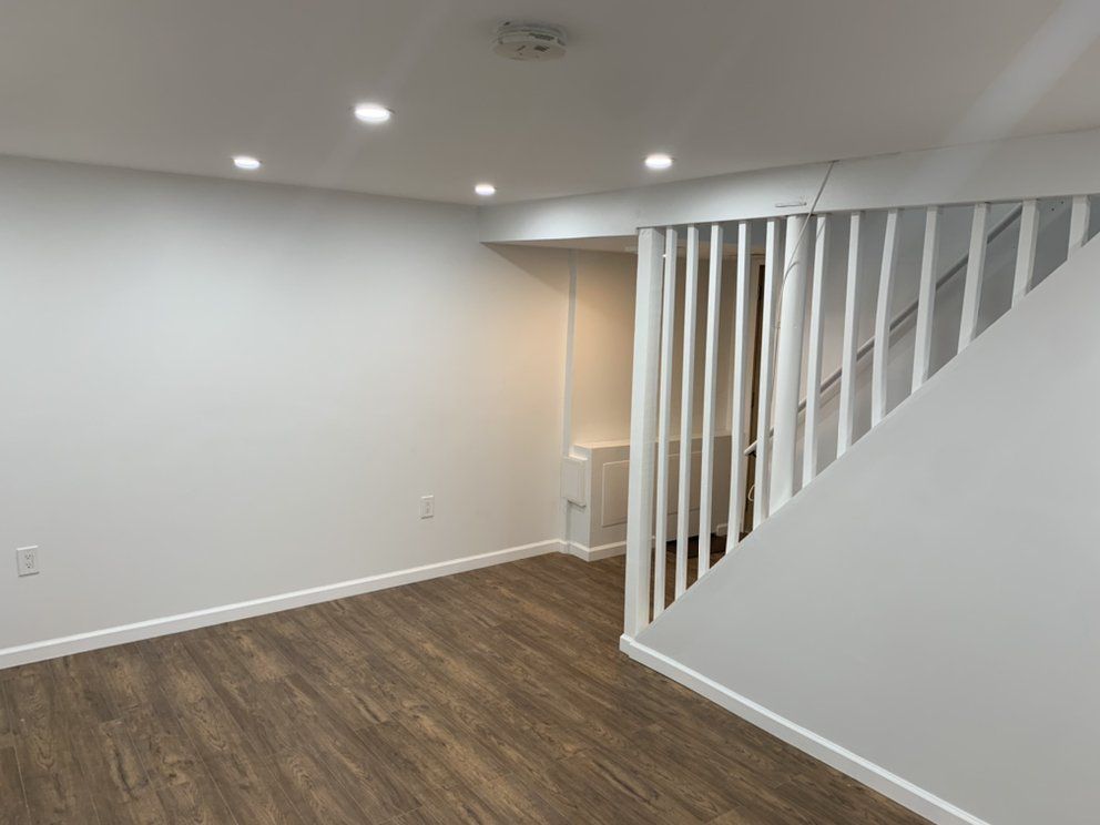 Empty room with wood-look floor, white walls, recessed lighting, and a partial staircase with vertical slats.