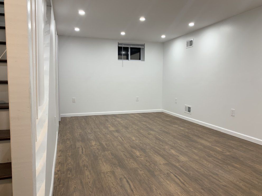 Empty, white-walled room with a small window, recessed lighting, and wood-look flooring. A staircase is visible.