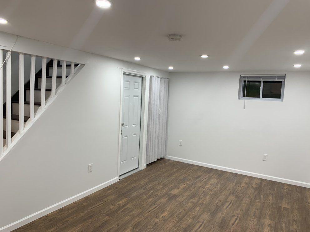 Basement interior with white walls, recessed lights, wooden floors, door, stairs, small window.