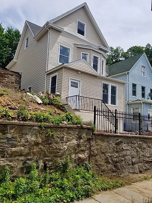Beige multi-story house on a hillside with stone retaining wall and black fence.