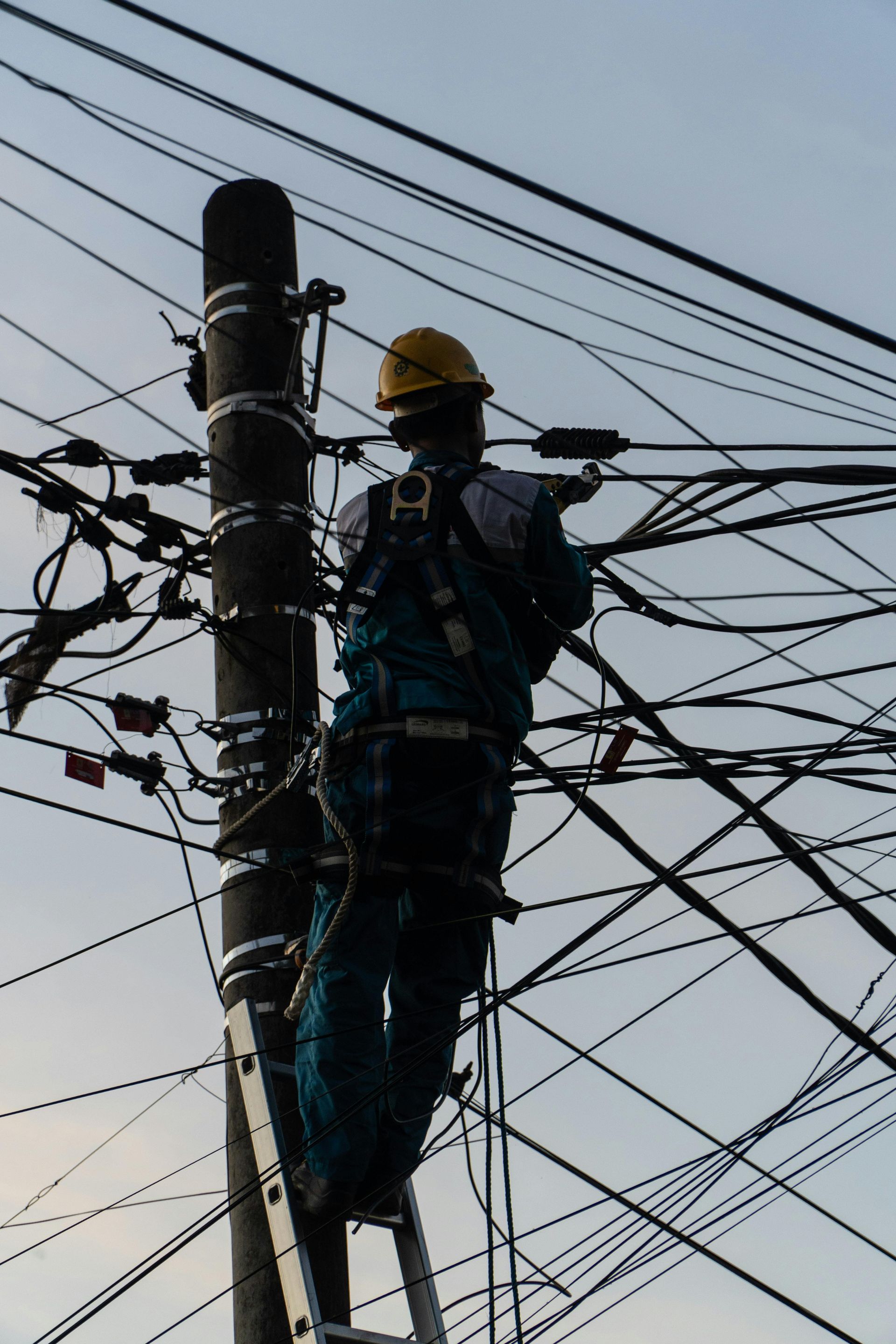 Lineman on ladder, working on a power pole with many wires, against a cloudy sky.