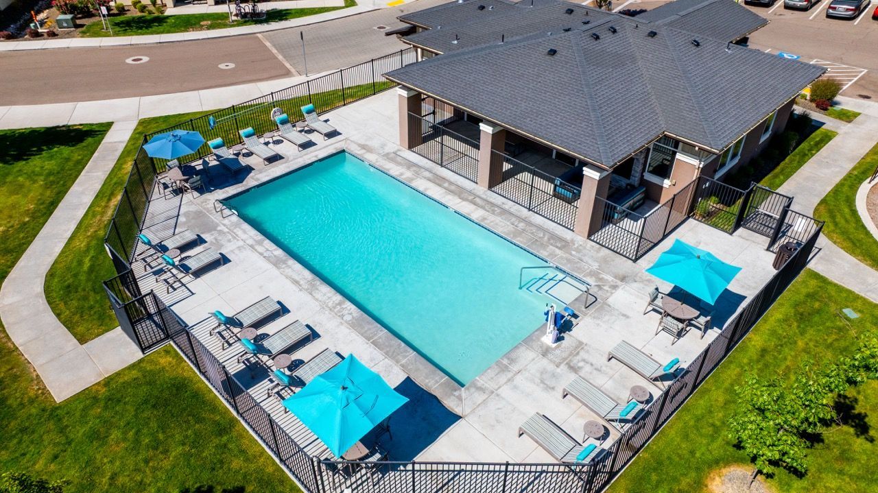 An aerial view of a fenced community pool with lounge chairs and blue umbrellas beside a clubhouse.