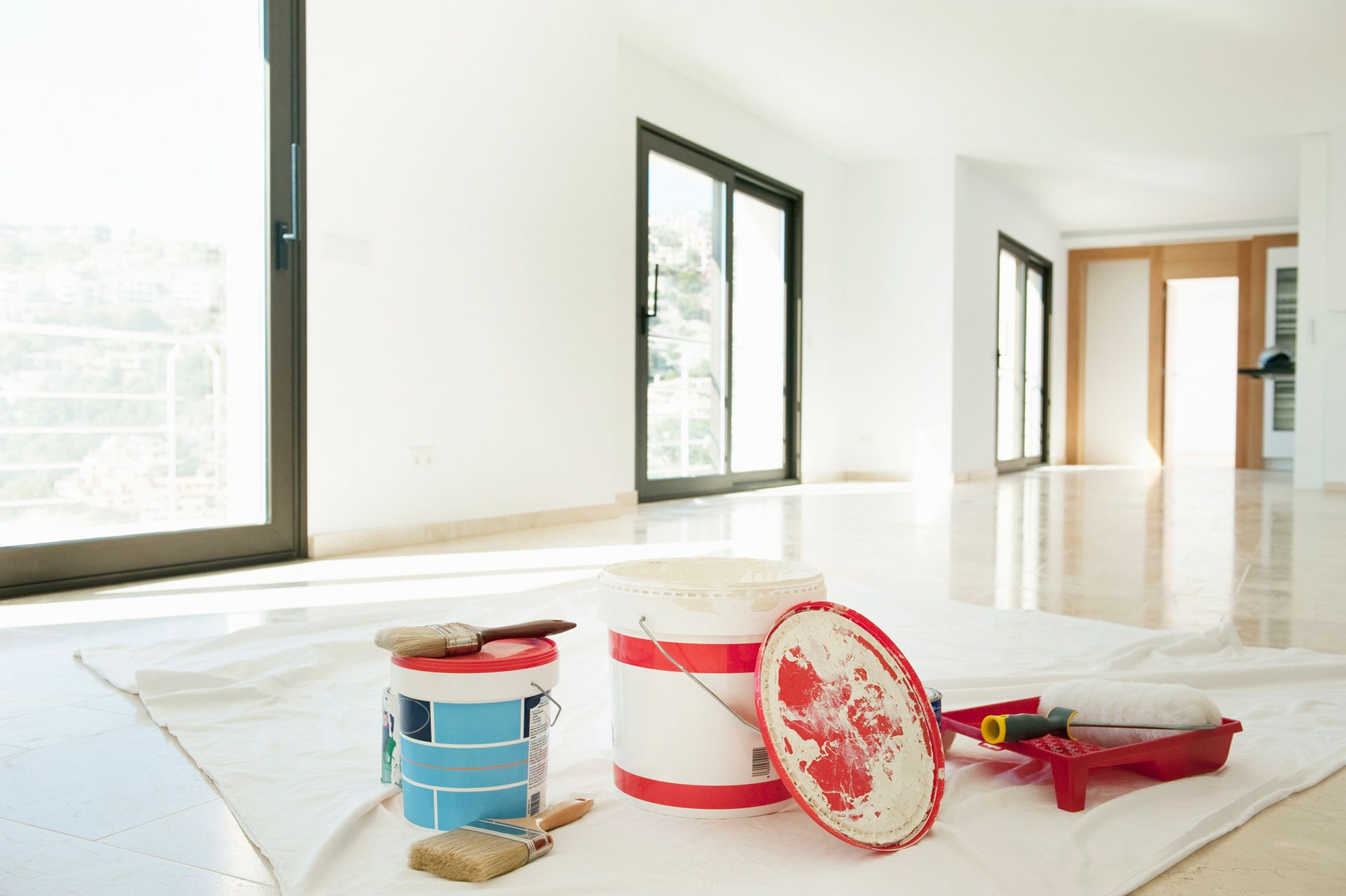 Three buckets of paint and brushes are sitting on a white towel in an empty room.