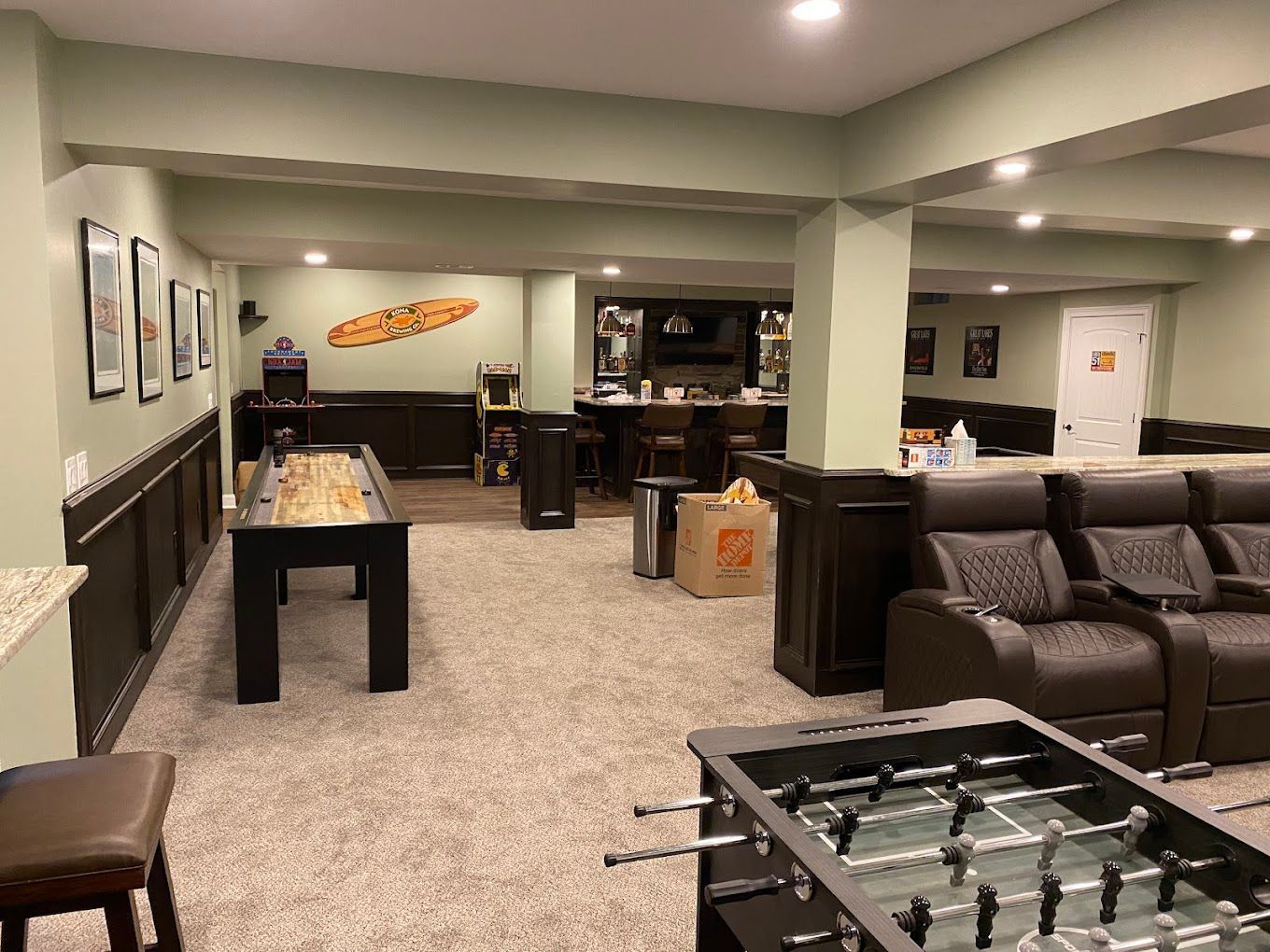 A finished basement features a shuffleboard table, foosball table, home theater seating, and a bar area with light green walls.
