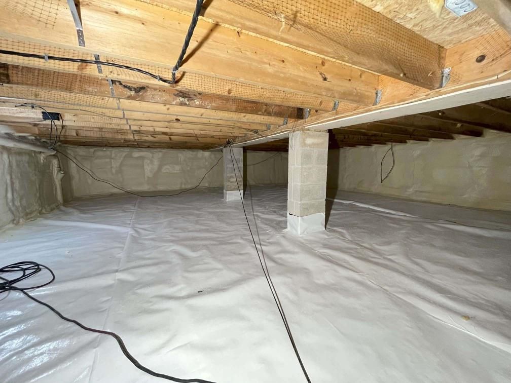 A crawl space with a clean white vapor barrier on the ground, concrete support pillars, and exposed wooden joists above.