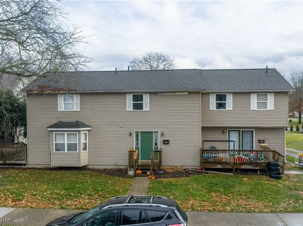 A beige, two-story house with a grey roof, a green front door, a bay window on the left, and a wooden deck on the right.