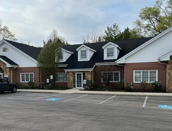 A brick commercial building with white gables, dormer windows, and a black shingled roof next to a paved parking lot.