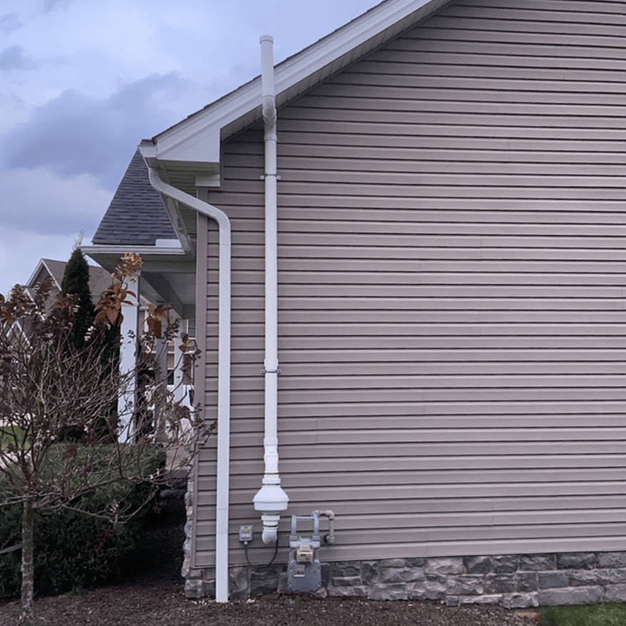 A vertical white radon exhaust pipe runs up the beige vinyl-sided exterior wall of a house near a gas meter.