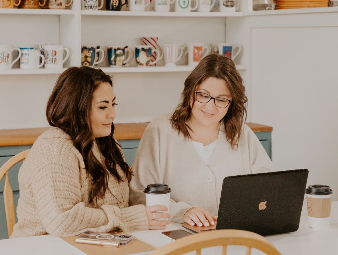 Two women are sitting at a table looking at a laptop