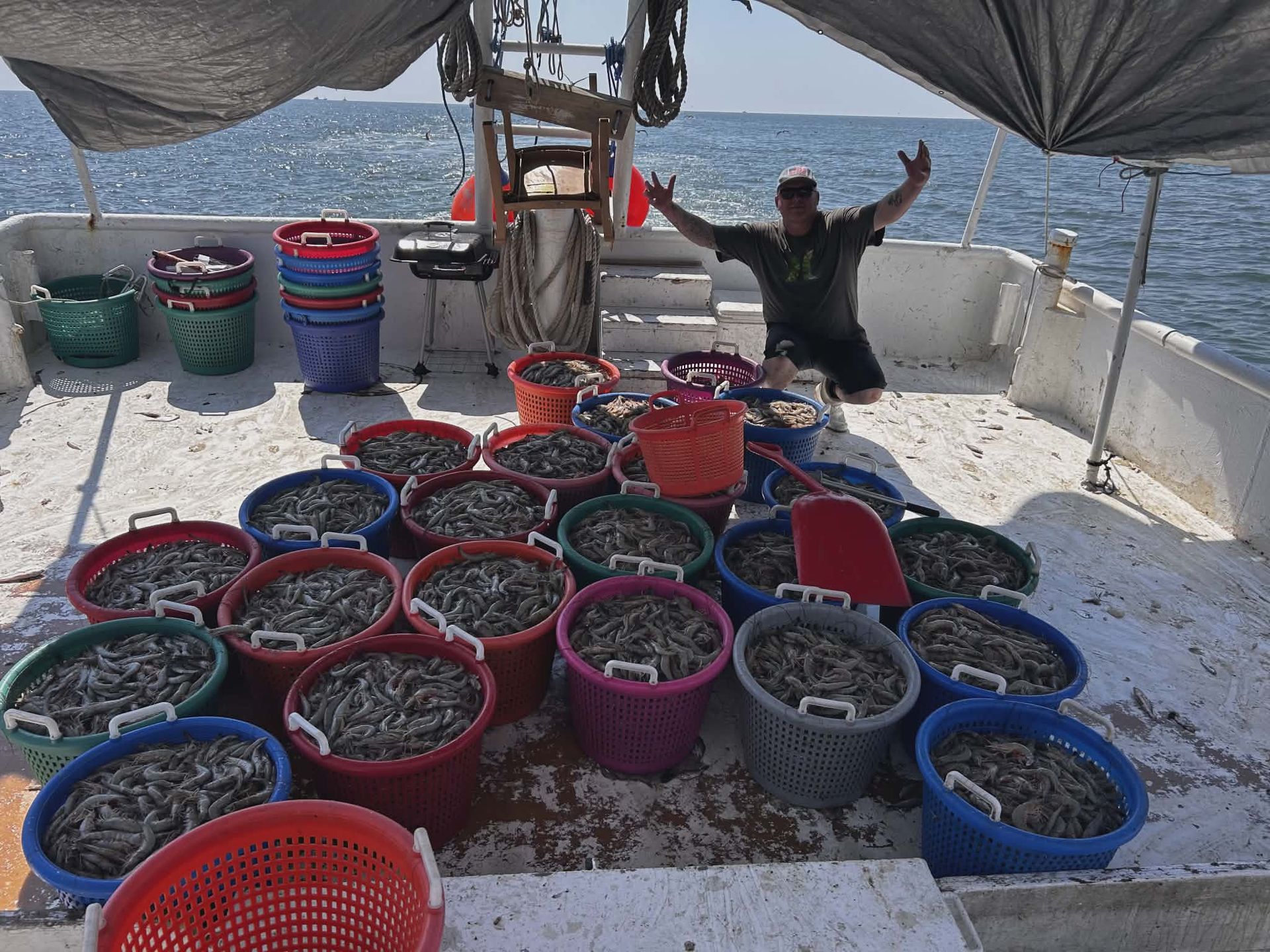 Man on boat surrounded by full colorful baskets of fish, arms raised, on a sunny day.